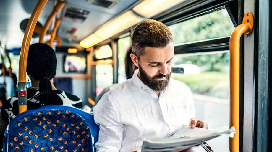A man is sitting on the bus. He is wearing a white crinkled shirt, and is reading the newspaper.