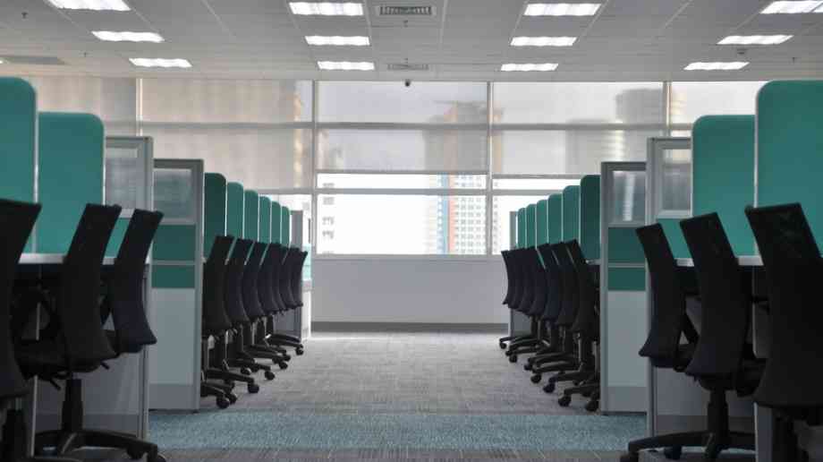 The picture shows a row of empty chairs and desks in an office. It is a well lit room filled with plenty of natural light.
